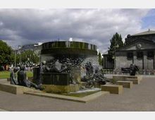 Ein runder Brunnen mit Bronzeskulpturen von Menschenfiguren, die um den Rand verteilt sind. Im Hintergrund sind ein historisches Gebäude mit Säulen und Bäume zu sehen, unter einem bewölkten Himmel.