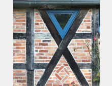 Fachwerkfassade mit rotem Backsteinmauerwerk und dunklen Holzbalken in einem Kreuzmuster, ergänzt durch ein blaues, dreieckiges Fenster. Im Vordergrund sind einige Pflanzen zu sehen.
