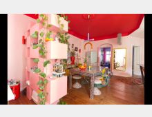 Ein bunter Innenraum in einem modernen Stil mit rosa Wänden und einer roten Decke. Zu sehen sind ein Regal mit Pflanzen, ein Esstisch mit Fliesenmuster und bunten Stühlen sowie diverse Dekorationselemente; im Hintergrund befinden sich ein großer Spiegel mit goldenem Rahmen und eine Küchenecke mit einem Retro-Kühlschrank.