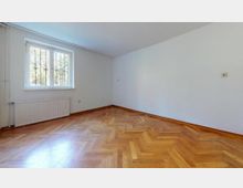 Ein leerer Raum mit hellem Fischgrätenparkettboden, weißen Wänden und einem Fenster mit Gitterstäben. Unter dem Fenster befindet sich ein Heizkörper, und die rechte Wand ist ohne weitere Gestaltungselemente.