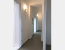 Ein minimalistischer, heller Flur mit weißen Wänden, grauem Boden und Wandlampen, die warmes Licht spenden. Die schlichte Architektur zeigt scharfe Linien und hohe Decken.