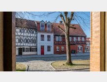 Straßenszene mit historischen Fachwerkhäusern, darunter ein weißes und ein rotes Gebäude, die sich entlang eines gepflasterten Gehwegs erstrecken. Im Vordergrund steht ein Baum mit kahlen Ästen, der den Blick auf die Häuser leicht verdeckt.