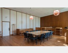 Konferenzraum mit schlichter Holzeinrichtung, bestehend aus einem großen rechteckigen Tisch mit blauen Polsterstühlen, Wandverkleidungen aus Holz und einem Parkettboden. An der Decke hängen zwei Designerleuchten, und im Hintergrund befinden sich Regale und ein Kleiderständer.