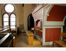 Innenraum einer Kirche mit einer kleinen roten Orgel an der rechten Wand. Im Vordergrund stehen Holzbänke und -stühle, links sind drei bunt verglaste Fenster mit einem runden oberen Fensterabschnitt zu sehen.