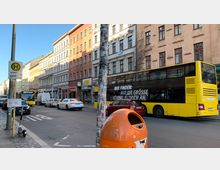 Straßenszene in einer Stadt mit mehrstöckigen Wohn- und Geschäftsgebäuden im Altbaustil; im Vordergrund stehen ein orangefarbener Mülleimer und ein Straßenschild mit Busfahrplan. Ein gelber Doppeldeckerbus sowie weitere Autos sind auf der Straße zu sehen.