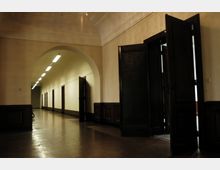 Flur in einem historischen Gebäude mit hohen Decken, einem großen Rundbogen und hölzernen Wandverkleidungen. Der Gang ist von mehreren Türen aus dunklem Holz und beleuchteten Deckenlampen gesäumt.