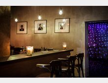 Eine kleine Bar mit dunkler Holztheke, darauf Kerzen und Getränkeflaschen. An der grauen Wand hängen gerahmte Porträtfotos, daneben führt eine Türöffnung in einen bunt beleuchteten Raum mit Lichtpunkten.
