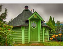 Kleines, achteckiges Holzgebäude mit grün gestrichenen Wänden und einem dunklen Spitzdach aus Schindeln, umgeben von Bäumen und Sträuchern. Der Eingang hat eine grüne Tür mit achteckigem Fenster, darüber befindet sich ein gelbes Schild mit Hinweistext.