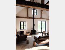 Wohnzimmer mit hohen Decken, sichtbaren Holzbalken und großen Fenstern, die viel Tageslicht hereinlassen. Zentral steht ein schwarzer Kaminofen, umgeben von Sofas und Holzstühlen; Holzscheite und Körbe mit Kaminholz sind dekorativ aufgestellt.