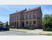 Ein zweistöckiges Gebäude aus rotem Backstein mit einem Satteldach und großen rechteckigen Fenstern. Vor dem Gebäude befinden sich ein Zaun, ein Gehweg aus Kopfsteinpflaster und ein geparktes Auto; daneben stehen Bäume und Büsche.