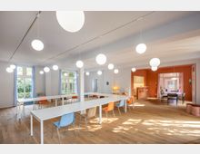 Heller Seminarraum mit weißen Wänden, großen Fenstern mit blauen Vorhängen und einem Holzfußboden. Lange weiße Tische sind in U-Form angeordnet, umgeben von bunten Stühlen; im Hintergrund befindet sich ein Bereich mit einer orangen Wand, einer Küche und einem weiteren Tisch.