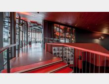 Moderne Innenansicht eines Gebäudes mit großen Glasfenstern, die viel Tageslicht hereinlassen. Der Raum ist mit roten Treppen, Holzdecken und schwarzen Elementen gestaltet; links befindet sich ein langer Gang mit Ausblick, rechts eine Wand mit Porträtfotos hinter einem Geländer.