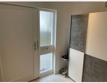 Eine helle Wohnungseingangstür mit einem seitlichen Fenster aus strukturiertem Glas. Rechts steht ein grauer und weißer Einbauschrank, darunter ein Regal mit Schuhen.