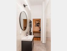 Schmales, modernes Badezimmer mit großformatigen weißen Marmorfliesen, grauem Boden und einem schwarzen Waschtischunterschrank mit rechteckigem Waschbecken und rundem Wandspiegel. An der Wand sind eine lineare LED-Leuchte und ein schwarzer Handtuchheizkörper zu sehen; am Ende öffnet sich der Raum zu einem Flur mit Holzkonsole und Wandbild.