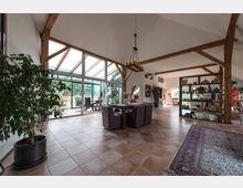 Großzügiger Wohnraum mit offenem Dachstuhl und Holzbalken, großen Glasflächen, die auf eine Terrasse mit Blick ins Grüne hinausführen. Auf dem Fliesenboden stehen ein Esstisch mit Stühlen, Zimmerpflanzen und ein Regal mit Dekoration.