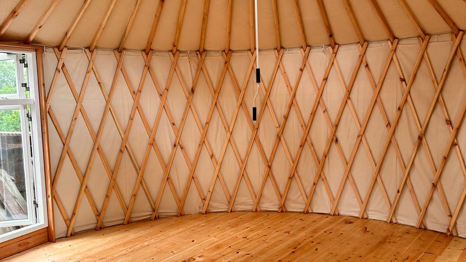 Innenansicht einer Jurte mit holzverkleidetem Boden, einer runden Dachstruktur aus Holzbalken und einem zentralen runden Dachfenster. Die Wände bestehen aus einem diagonalen Holzrahmen, der mit hellem Stoff bespannt ist.