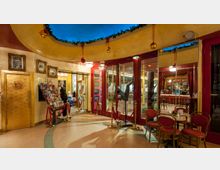 Eingangsbereich eines eleganten Theaters oder Varietés mit goldenen und roten Designelementen. Zu sehen sind ein goldener Türrahmen mit der Aufschrift "Wintergarten", rote Stühle und Tische, dekorative Beleuchtung, sowie Schaufensterpuppen und ein Blick in eine Bar mit klassischem Holzinterieur.