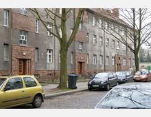 Straßenansicht eines Wohngebäudes mit grauer Fassade und Backsteinelementen an Eingängen und Fenstern. Vor dem Gebäude stehen Bäume, geparkte Autos und eine Abfalltonne, auf einem Kopfsteinpflasterbelag.