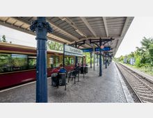 Ein S-Bahnhof mit dem Schild „Yorckstraße“, überdacht durch ein Metallgitterdach, getragen von verzierten, dunkelblauen Säulen. Auf der linken Seite fährt eine gelb-rote S-Bahn ein, während sich rechts die Gleise und eine grüne, bewachsene Umgebung erstrecken.