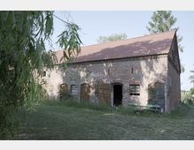 Altes Backsteingebäude mit rotem Ziegeldach und mehreren Fenster- und Türöffnungen, teilweise mit Holztüren oder Brettern verschlossen. Im Vordergrund eine Wiese, links von einem herabhängenden Baumzweig eingerahmt.