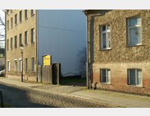 Ansicht einer Straßenecke mit zwei älteren Wohngebäuden, eines mit verputzter Fassade und das andere mit unverputztem Ziegelmauerwerk. Zwischen den Gebäuden befindet sich ein Holztor mit einem Schild, das eine Tischlerei bewirbt; der Bodenbelag besteht aus Kopfsteinpflaster.