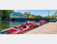 Roter Motorboothafen an einem Holzsteg mit einer blauen Wellblechmauer im Hintergrund, daneben ein Gebäude mit Werbeschildern für Räucherfisch, Fischbrötchen und Getränke. Im Hintergrund sind Bäume und Palmen zu sehen.