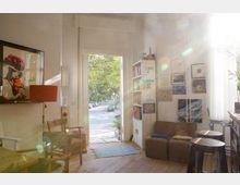 Helles Wohnzimmer mit Holzboden, einer offenen Tür, die auf eine von Bäumen gesäumte Straße führt. Die Wände sind mit Kunstwerken dekoriert, ein Bücherregal steht neben der Tür, und verschiedene Sitzgelegenheiten sowie Möbel wie Stühle und Hocker sind im Raum angeordnet.