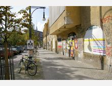 Straßenszene mit einem verwitterten Gebäude, dessen Fassade mit Graffiti und bunt bedruckten Plakaten bedeckt ist. Entlang des Gehwegs stehen geparkte Fahrräder und Bäume, im Hintergrund ist der Berliner Fernsehturm erkennbar.
