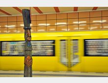 Holzskulptur in einer U-Bahn-Station mit gelben Kacheln an der Wand und einer vorbeifahrenden gelben U-Bahn im Hintergrund. Die Skulptur zeigt geschnitzte, farbig bemalte Figuren auf einem vertikalen Holzpfahl.