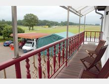 Blick von einem Balkon mit roten, verzierten Geländern und Holzboden auf einen Hof mit geparkten Fahrzeugen, einem Carport mit Wellblechdach und einer grünen Plane, umgeben von Wiesen und Bäumen im Hintergrund.