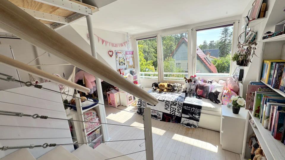 Helles Dachgeschoss-Kinderzimmer mit weißem Holzfußboden, großem Fensterband und Tagesbett unter den Fenstern. Im Vordergrund führt eine offene Treppe mit Holzhandlauf und Seilgeländer in den Raum; rechts stehen Regale mit Büchern und Dekoration.
