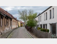 Schmale gepflasterte Straße in einer Wohnsiedlung mit alten und renovierten Häusern; links ältere Gebäude mit Putzschäden und Dachziegeln, rechts moderne Häuser mit hellen Fassaden und Holzjalousien. Bäume und Sträucher säumen den Weg, ein Holzzaun und ein abgestelltes Fahrrad sind sichtbar.