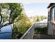 Blick von einem kleinen Balkon mit Metallgeländer an einem Backsteingebäude auf einen Fluss oder See. Im Vordergrund stehen hohe Bäume mit dichtem Laub, dahinter sind Ufervegetation, ein flaches Dach und entfernte Gebäude zu sehen.
