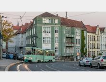 Straßenszene in einer Stadt mit einer grünen Straßenbahn, die um eine Kurve fährt, umgeben von mehrstöckigen Wohnhäusern mit historischen Fassaden. Im Vordergrund parkende und fahrende Autos, sowie Oberleitungen für die Straßenbahn.