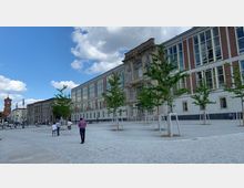 Ein offener städtischer Platz mit gepflastertem Boden, jungen Bäumen in Reihen und einem großen Gebäudekomplex mit klassizistischen und modernen Elementen. Im Hintergrund sind weitere historische Gebäude sowie der Turm des Roten Rathauses sichtbar, umgeben von einem teils bewölkten Himmel.