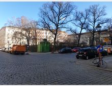 Straßenszene mit gepflasterter Kreuzung, parkenden Autos und einem grünen, historischen Pissoir aus Gusseisen. Im Hintergrund stehen Altbau-Wohnhäuser mit heller Fassade und kahlen Bäumen, Straßenschilder und ein Fahrrad sind am rechten Bildrand sichtbar.