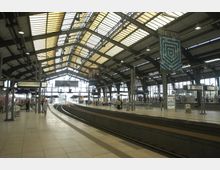 Eine überdachte Bahnhofshalle mit mehreren Bahnsteigen und Gleisen, umgeben von großen Glasfenstern und einem Dach aus Stahlträgern und lichtdurchlässigen Elementen. Menschen bewegen sich entlang der Bahnsteige, und Informationstafeln sind sichtbar.