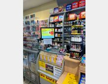 Ein kleiner Kiosk mit einem Verkaufsbereich für Tabakwaren, E-Zigaretten, und Lotto-Produkte, inklusive Regalflächen mit Zigarettendosen, einer Lottomaschine und Werbedisplays. Im Vordergrund liegen Lotto-Scheine, Werbeposter und kleine Verkaufsartikel.