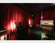 Small performance room with a low stage in front of red velvet curtains, lit by colored spotlights on a dark ceiling. A white upright piano, an acoustic guitar on a stand, stacked chairs, and audio equipment sit along the sides on a wooden floor.