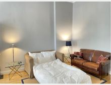 Wohnzimmer mit neutralen grauen Wänden, einem ausziehbaren beigen Sofa, das als Bett hergerichtet ist, einer braunen Ledercouch mit gemusterten Kissen und zwei beidseitig platzierten Nachttischen mit Lampen. Der Boden ist aus hellem Holz, und ein kleiner Glastisch steht neben dem Sofa.