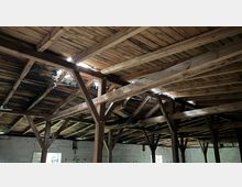 Holzinterieur einer alten, verlassenen Halle mit sichtbarer Dachkonstruktion. Das Dach weist beschädigte Stellen auf, durch die Licht einfällt, und die Wände bestehen aus ungepflegtem Mauerwerk.