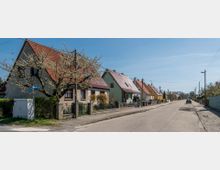 Wohnstraße mit einer Reihe von Einfamilienhäusern, die jeweils rote oder farbige Satteldächer haben, flankiert von Vorgärten mit blühenden Bäumen und niedrigen Zäunen. Die Szenerie zeigt eine asphaltierte Straße mit Gehwegen und vereinzelten Straßenlampen unter einem blauen Himmel.