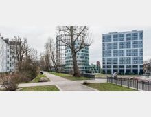 Ein urbaner Park mit Gehwegen und Rasenflächen, umgeben von moderner und historischer Architektur. Im Hintergrund sind ein runder, grün-weißer Büroturm, ein weißes mehrstöckiges Gebäude mit großen Fenstern und ein alter Gasspeicher zu sehen.