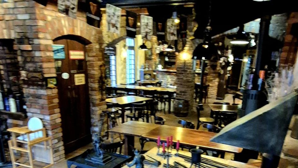 Rustikales Restaurantinterieur mit Backsteinwänden, Gewölbebögen und sichtbaren Holzbalken an der Decke. Mehrere Holztische und Stühle stehen entlang eines langen Raums, ergänzt durch hängende Lampen und dekorative Metallobjekte auf einem gemauerten Tresen.