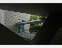 Außenansicht eines modernen Gebäudes mit einer Fassade aus hellen Putzwänden und regelmäßigen Punkten, daneben ein Vordach, das von Betonträgern gestützt wird. Im Hintergrund sind Bäume und Sträucher zu sehen.