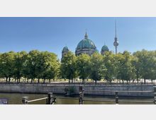 Blick auf den Berliner Dom mit seiner grünen Kuppel, umgeben von Bäumen, und im Hintergrund der Berliner Fernsehturm. Im Vordergrund verläuft ein Fluss mit einer Steinmauer am Ufer.