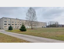 Mehrstöckiges, graues Plattenbaugebäude mit zahlreichen Fenstern, umgeben von einer großen Grünfläche und einigen kleinen Bäumen. Im Vordergrund verläuft ein asphaltierter Gehweg, seitlich steht ein Transporter sowie ein Auto, während im Hintergrund Bäume und Stromleitungen zu sehen sind.