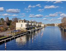 Reihenhäuser am Ufer eines Kanals mit Anlegestellen für Boote, umgeben von Bäumen und weiteren Gebäuden im Hintergrund unter einem blauen Himmel mit weißen Wolken.