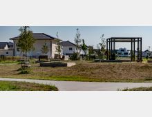 Kleiner Park in einem Wohngebiet mit jungen Bäumen, Bänken und einer offenen Pergola-Struktur. Im Hintergrund sind moderne Einfamilienhäuser mit dunklen Ziegeldächern sichtbar.