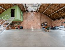 Großer Loft- oder Atelierraum mit Backsteinwänden, sichtbarem Holzdachstuhl und einem zentralen Oberlicht. Links hängt ein grünes Kubus-Zimmer mit Metalltreppe über einem Bücherregal, rechts stehen ein Esstisch mit Stühlen und ein Sofa vor einem bogenförmigen Fenster.